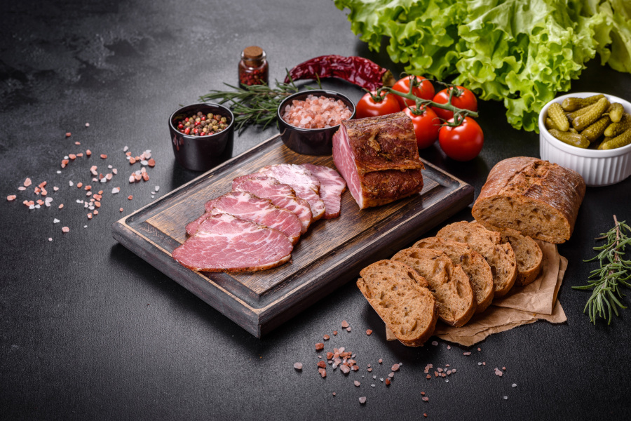 Aufgeschnittenes Fleisch und Brot auf einem Brett, dazu Tomaten, Gurken und Gewürze.