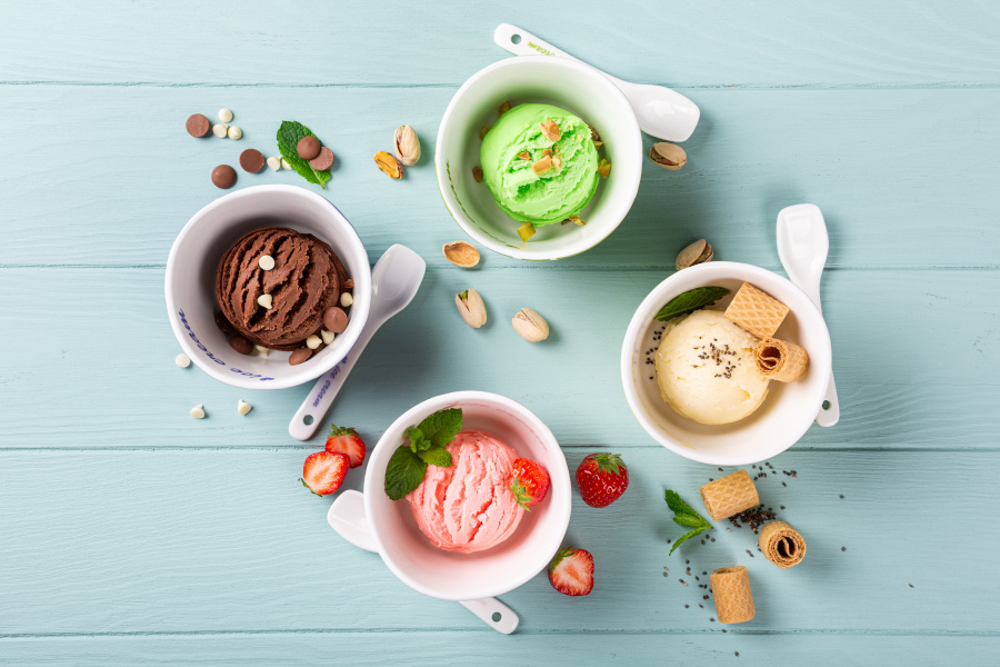 Drei Tassen mit verschiedenen Eiskugeln und Toppings auf einem hellblauen Tisch.