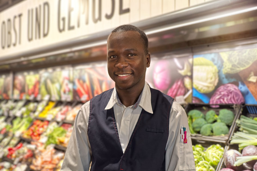 Mitarbeiter im Supermarkt vor der Obst- und Gemüseabteilung, lächelnd vor den Regalen.