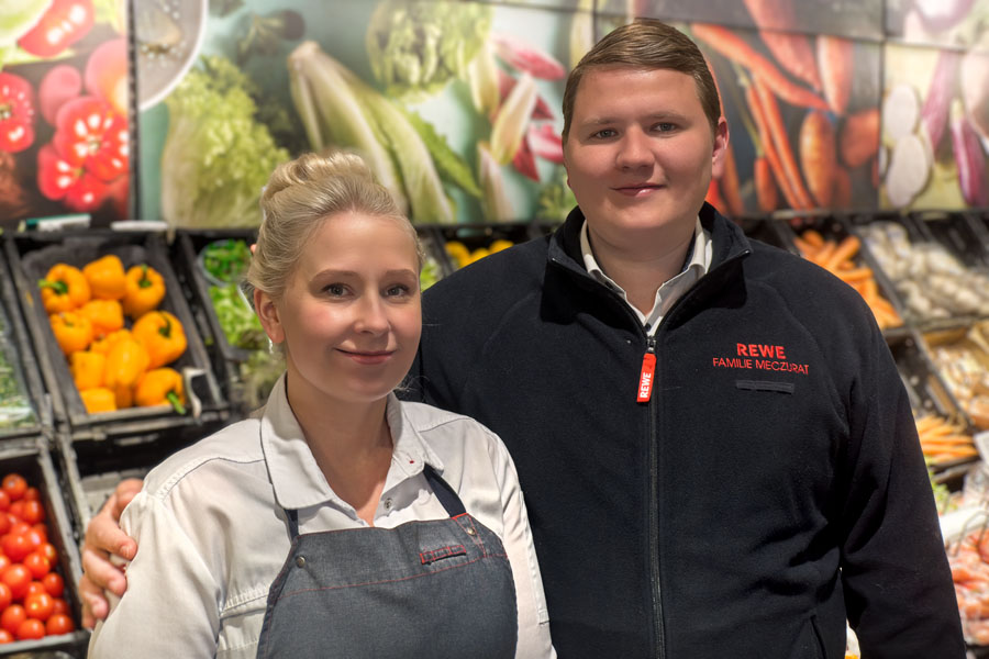 Zwei Supermarktmitarbeiter stehen vor der Obst- und Gemüseabteilung und schauen in die Kamera.