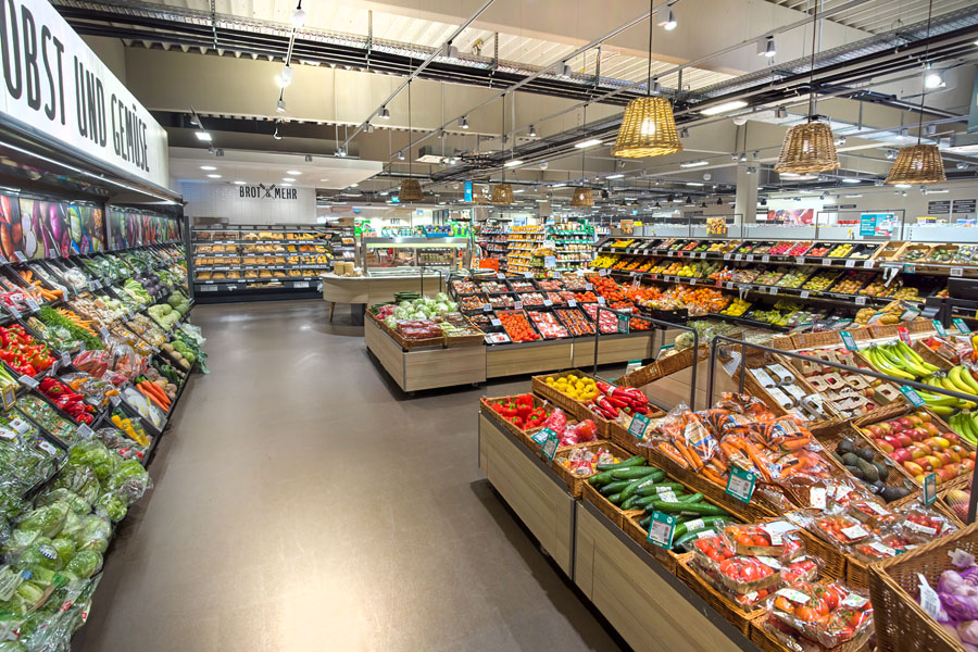 Große Obst- und Gemüseabteilung im Supermarkt mit vielen frischen Produkten und Brottheke im Hintergrund.